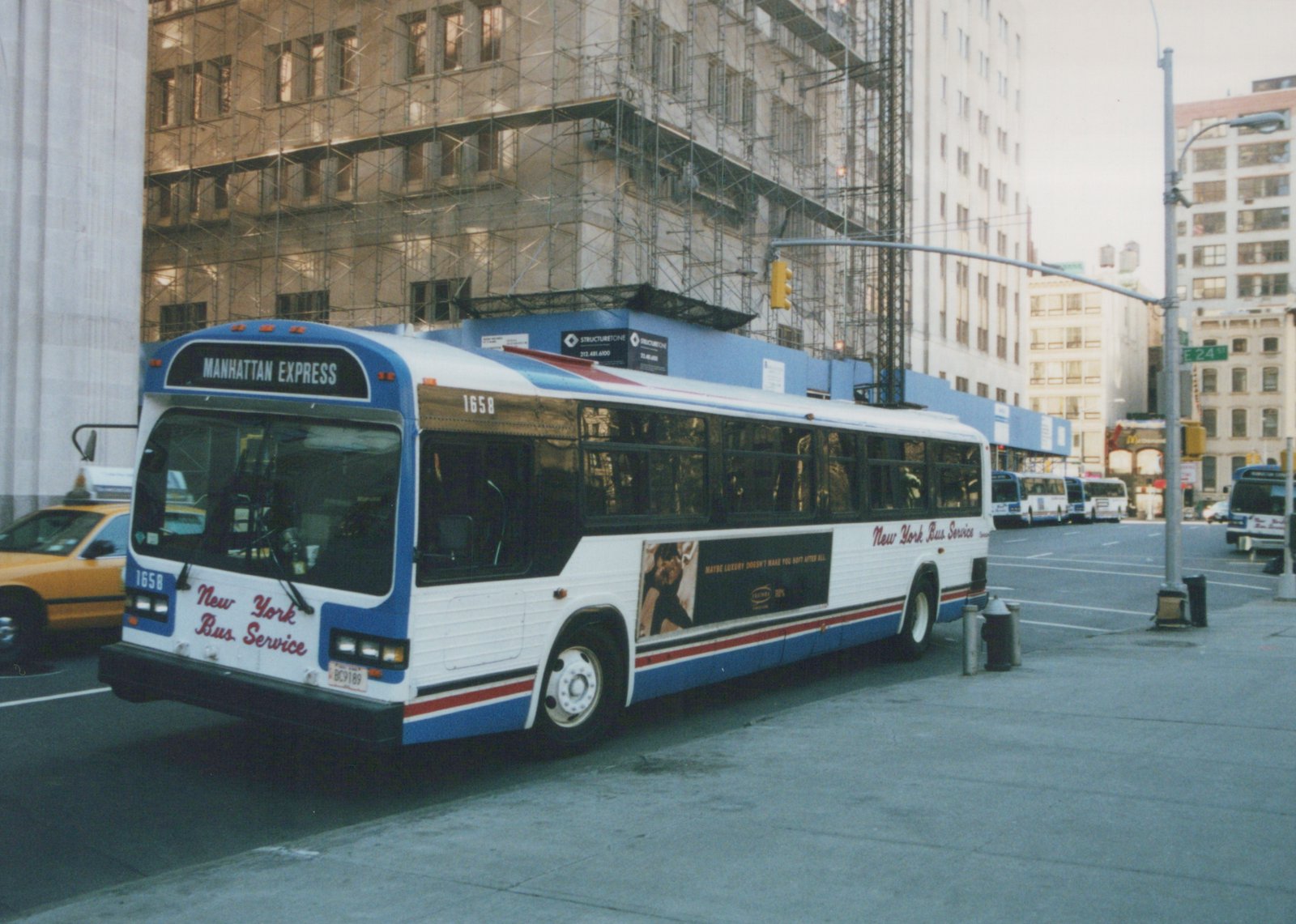 A white, blue and red New York Bus Service bus, numbered 1658 and marked 'Manhattan Express', is parked on a city street next to a sidewalk, with other buses and a yellow taxi behind it and tall buildings in the background.