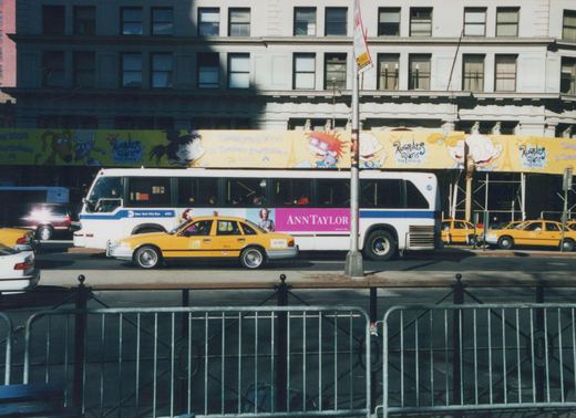 A white and blue MTA bus, number 4151, is driving down a city street past yellow taxis with a large 'Rugrats Paris THE MOVIE' billboard above it.