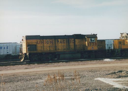 A side view of a yellow Union Pacific locomotive, number 1738, parked on railroad tracks in a gravel yard with patches of snow on the ground.