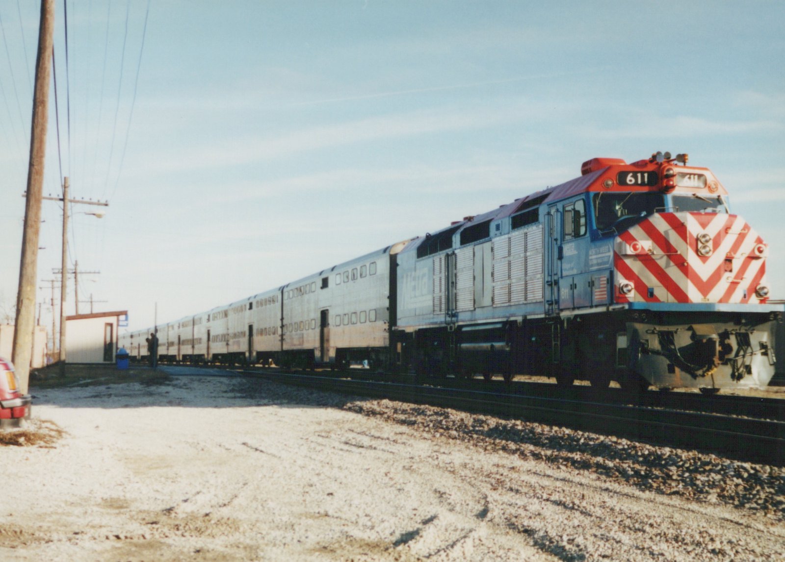 A blue and red Metra diesel locomotive with the number 611 pulls a line of silver double-decker commuter cars on a railroad track next to a small station platform with snow on the ground.
