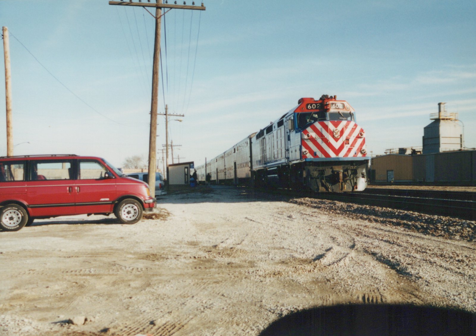 A red and silver Metra locomotive numbered 602 is parked on tracks next to a gravel lot with a red minivan. People stand near the train cars and buildings in the background under a clear sky.