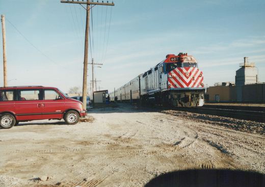A red and silver Metra locomotive numbered 602 is parked on tracks next to a gravel lot with a red minivan. People stand near the train cars and buildings in the background under a clear sky.