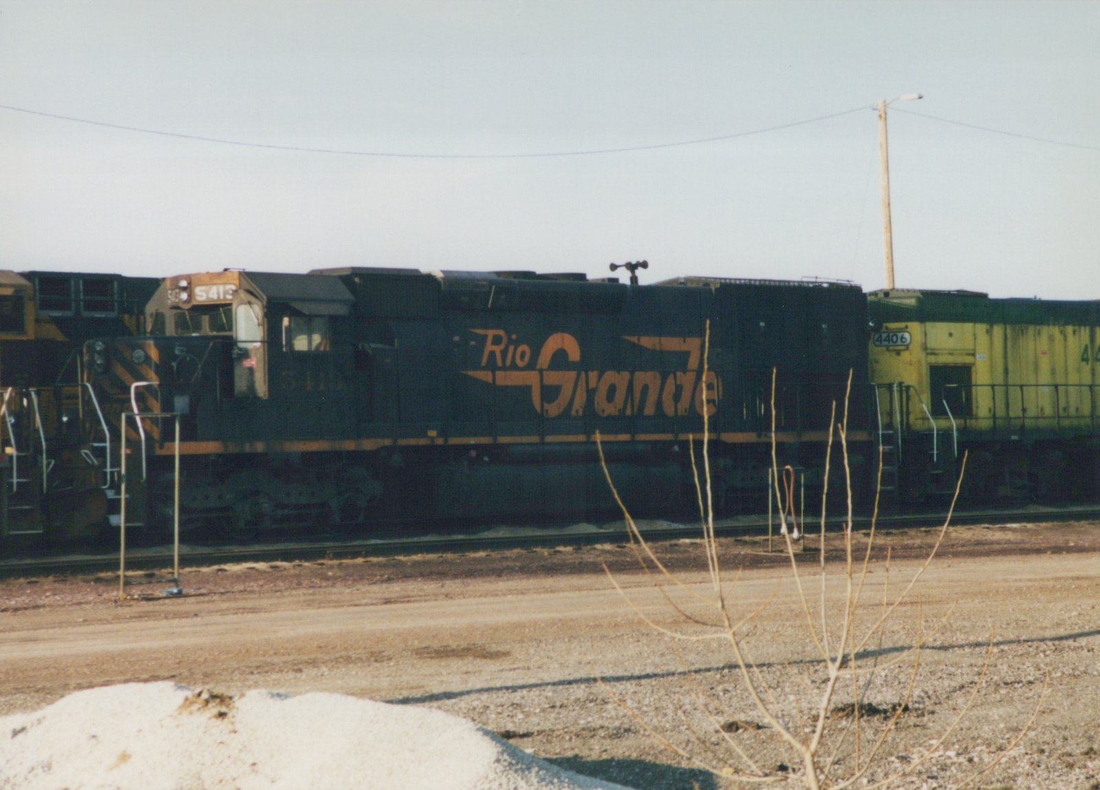 Side view of a dark green Rio Grande diesel locomotive, number 5413, with a yellow locomotive, number 4406, behind it, in a gravel yard under an overcast sky.