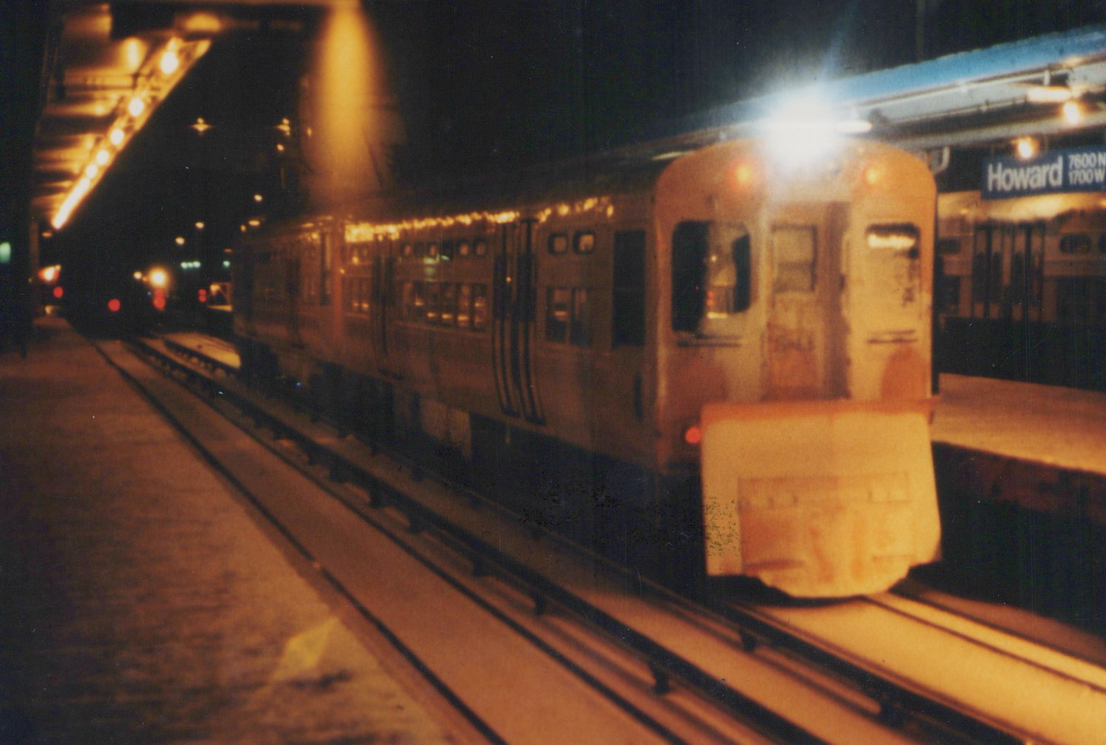 A silver rapid transit train with black stripes is stopped at a station platform at night, with the sign 'Howard 7600 N 1700 W' visible.