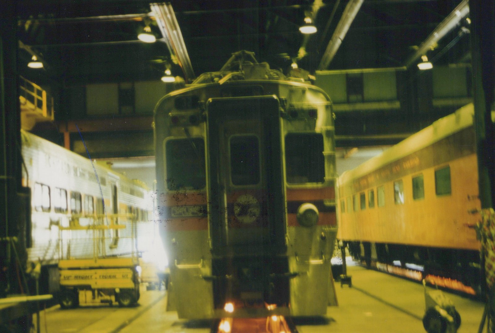 Rear view of a South Shore Line electric multiple-unit car inside a rail maintenance shop, with other cars and a yellow utility vehicle visible.