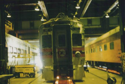 Rear view of a South Shore Line electric multiple-unit car inside a rail maintenance shop, with other cars and a yellow utility vehicle visible.