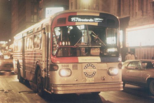Front three-quarter view of a red, white, and blue Chicago Transit Authority bus, number 3444, on a city street at night with its headlights on and a driver visible.