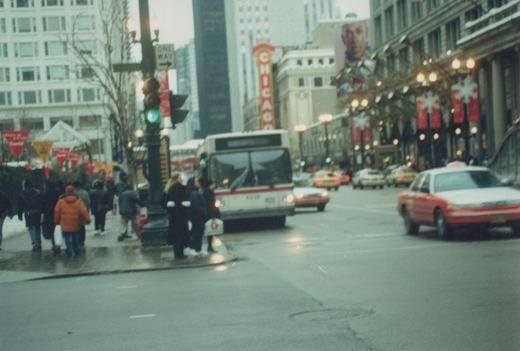 A white and red CTA bus with the number 4228 on its front, stopped on a wet city street lined with buildings and decorated for the holidays, with pedestrians on the sidewalk and cars in traffic.