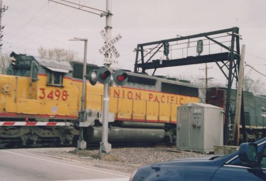 A yellow Union Pacific diesel locomotive numbered 3498 is moving across a street at a grade crossing, with flashing red lights and a railroad crossing sign visible.