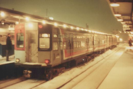 A long silver and red train sits at a snowy, elevated station platform at dusk, with a few people standing nearby.
