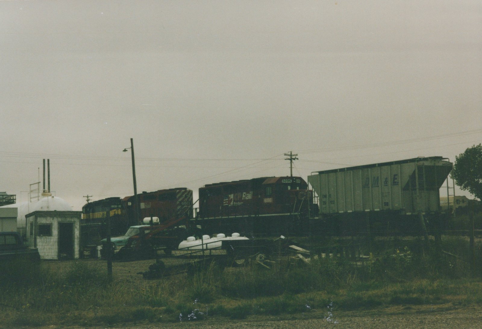 A side view of a Canadian Pacific diesel locomotive with red and black paint, numbered 6096, pulling a gray freight car with DM&E lettering, in a grassy rail yard with utility poles and overcast skies.
