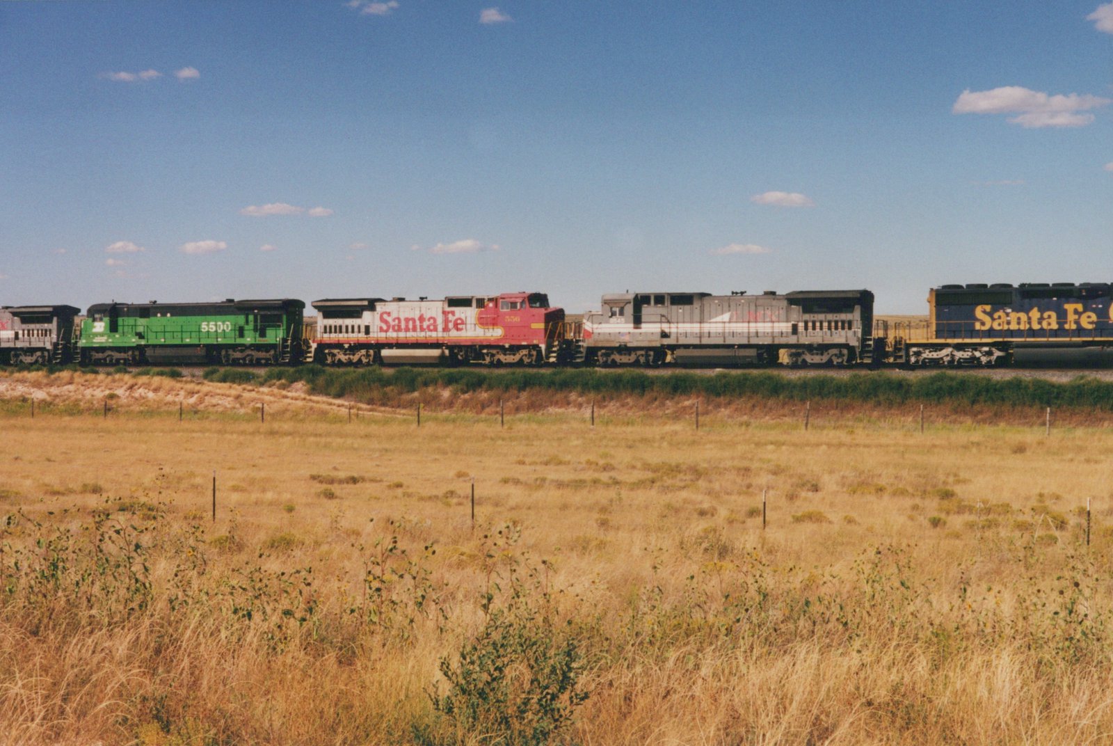 A line of four Santa Fe diesel locomotives in different paint schemes—green, red and white, silver, and blue—are on a railroad track beside a dry, grassy field under a clear blue sky.