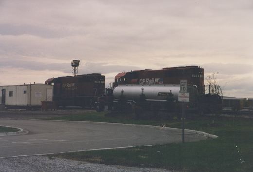 A brown CP Rail locomotive numbered 778 stands in a yard beside a white tanker truck and a portable building, with a speed limit sign in the foreground under a cloudy sky.