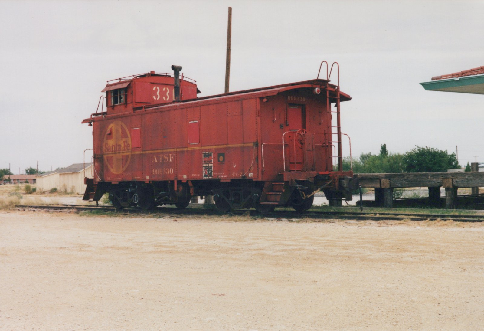 A red Santa Fe caboose, number 999330, viewed from a three-quarter rear angle, sits on a gravel track with a wooden platform in the background under an overcast sky.