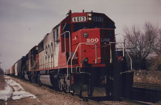 A red, white and black SOO Line diesel locomotive, number 6013, is shown pulling a long line of freight cars along a snow-dusted railroad track under an overcast sky, with bare trees in the background.