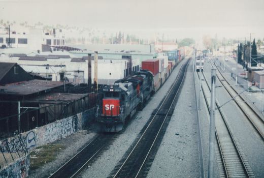 A black and red Southern Pacific locomotive numbered 8013 pulls a freight train on one set of tracks, while a white and red rapid transit car runs on electrified tracks nearby, under an overcast sky.