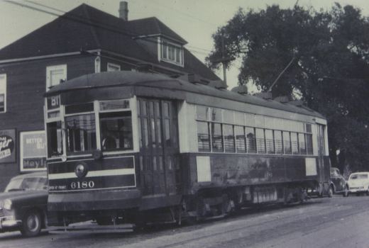 A black and white photograph of a vintage Chicago Surface Lines streetcar numbered 6180, showing its side and front with a trolley pole raised, parked on a street with cars and a building in the background.
