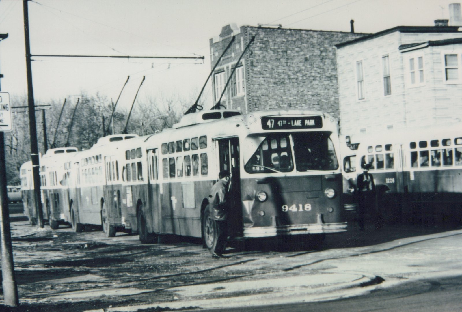 A black and white photo of several CTA trolleybuses lined up on a street in winter; the lead bus, number 9418, has its destination sign reading '47 47th - LAKE PARK' and a passenger is boarding.