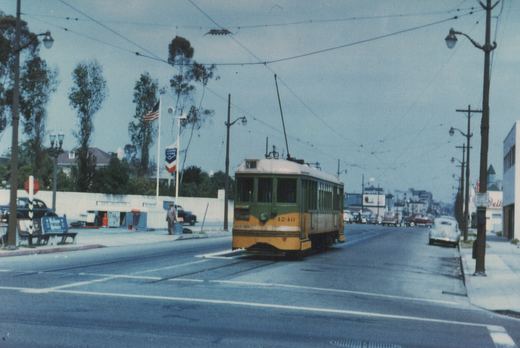 A green and yellow PCC streetcar numbered 1240 travels down a street lined with 1950s automobiles, overhead wires, and storefronts.
