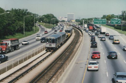 A CTA rapid transit train travels on tracks in the median of a multi-lane highway, surrounded by cars and trucks under a clear sky.