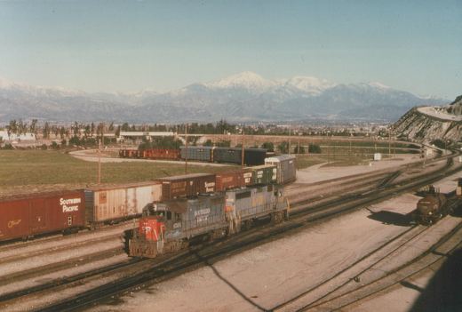 A Southern Pacific GP35 locomotive, number 6564, pulling a line of freight cars through a railroad yard with snow-capped mountains visible behind it.