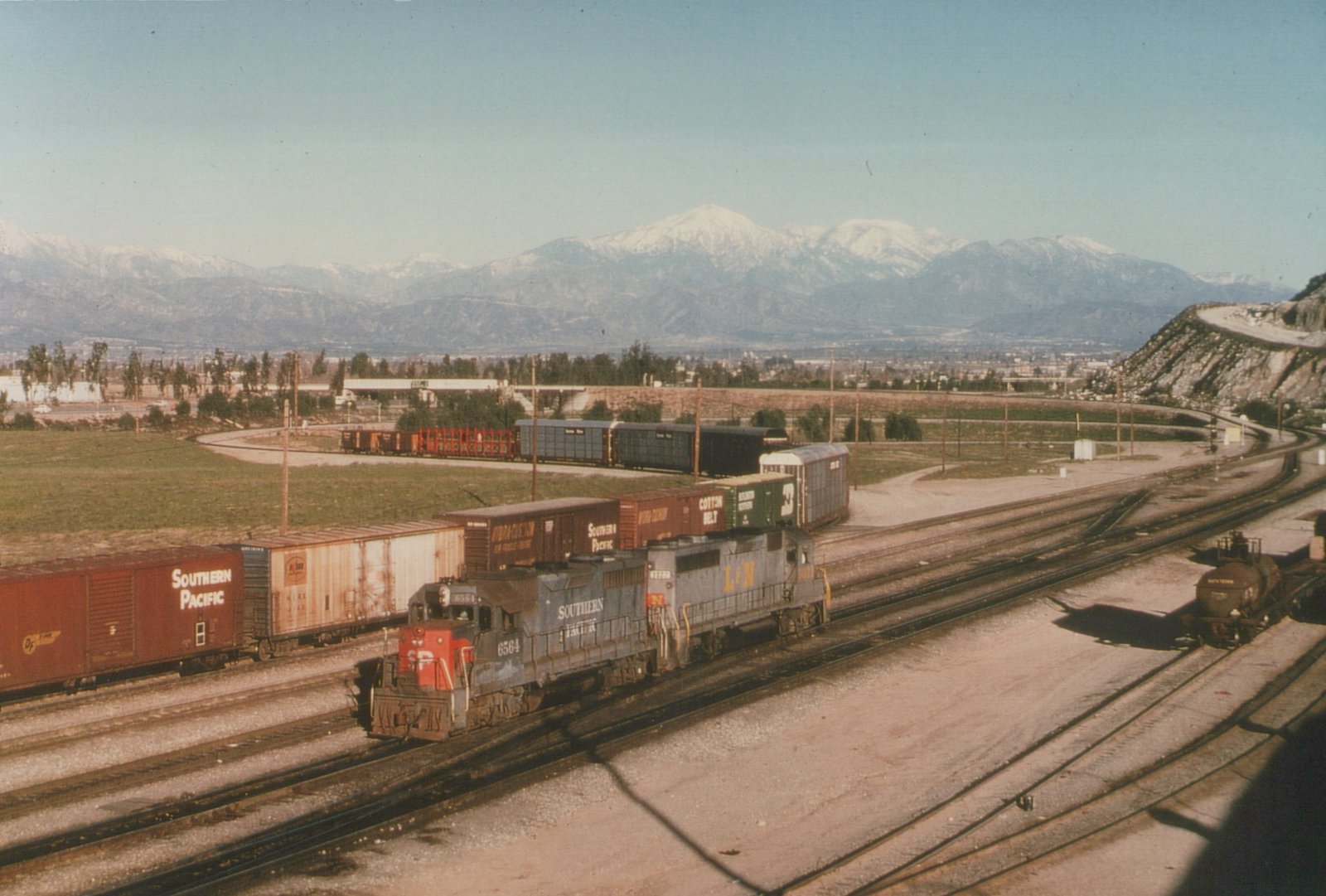 A Southern Pacific GP35 locomotive, number 6564, pulling a line of freight cars through a railroad yard with snow-capped mountains visible behind it.