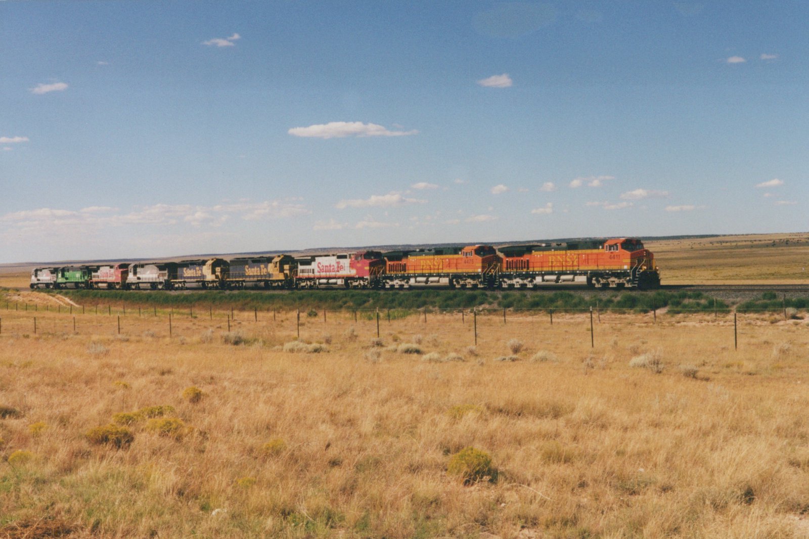 A long freight train with BNSF and Santa Fe locomotives runs on a railroad main line through an open, dry grassland under a blue sky with scattered clouds.