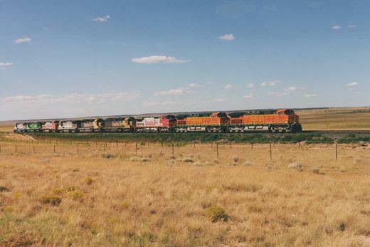 A long freight train with BNSF and Santa Fe locomotives runs on a railroad main line through an open, dry grassland under a blue sky with scattered clouds.