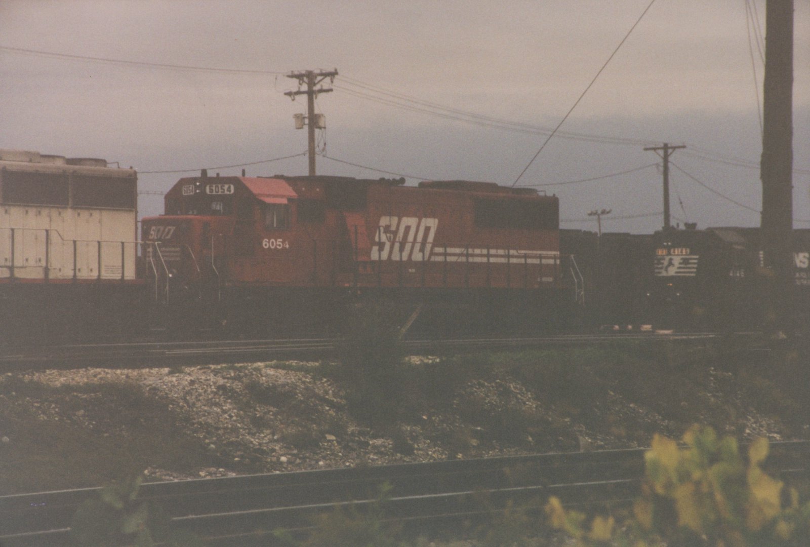 A red Soo Line locomotive with the number 6054 is in the foreground, leading a train, while a black Norfolk Southern locomotive with the number 4146 is visible behind it on an overcast day.