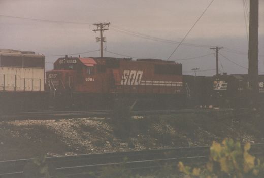 A red Soo Line locomotive with the number 6054 is in the foreground, leading a train, while a black Norfolk Southern locomotive with the number 4146 is visible behind it on an overcast day.