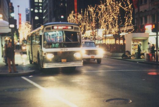 A white and dark-colored bus with the number 3679 on its front, operating on route 121, drives down a city street at dusk with string lights on the trees. Pedestrians are on the sidewalk, and another white vehicle is nearby.