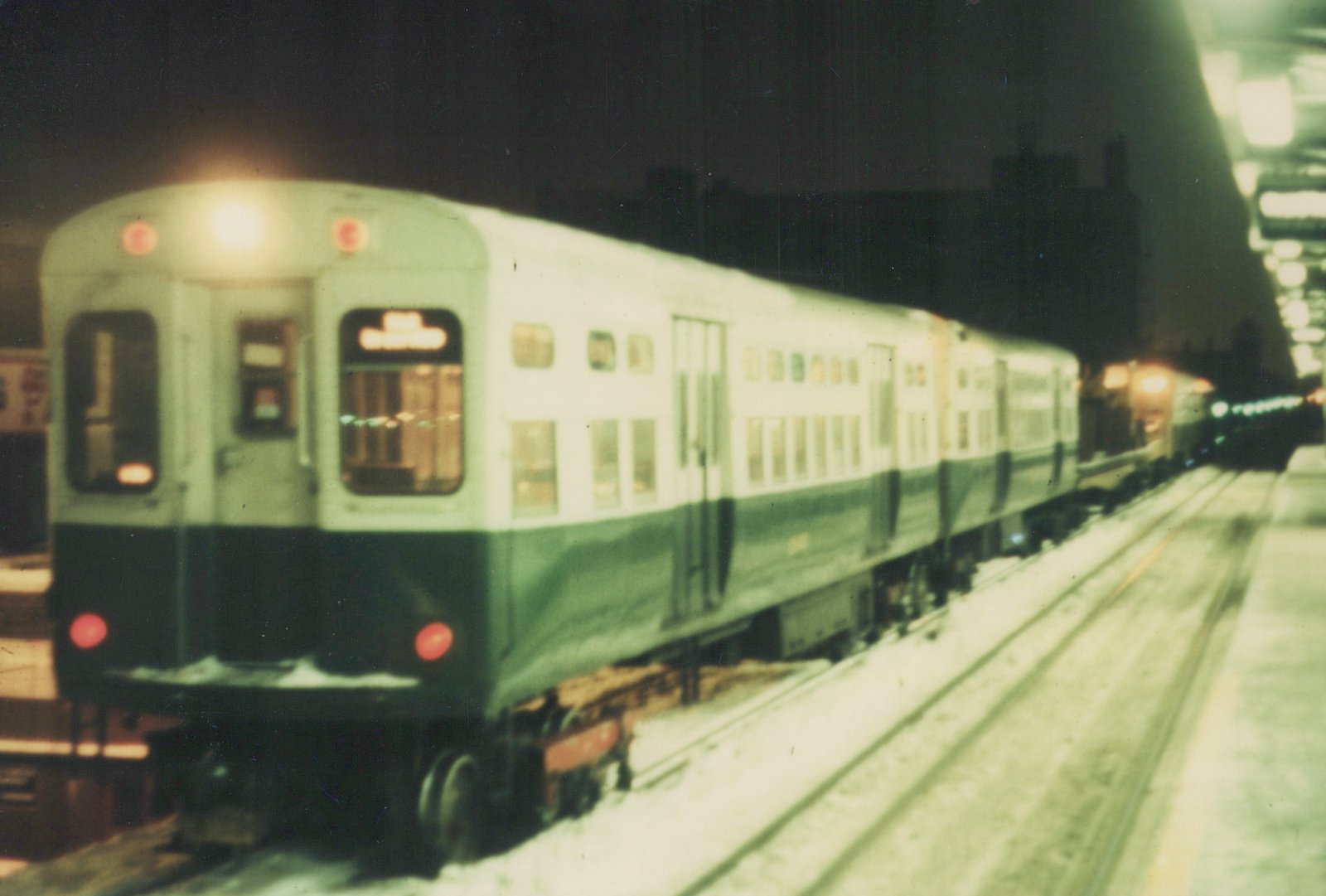 A blurry image of a train at night on an elevated track with snow on the ground and platform, viewed from behind.