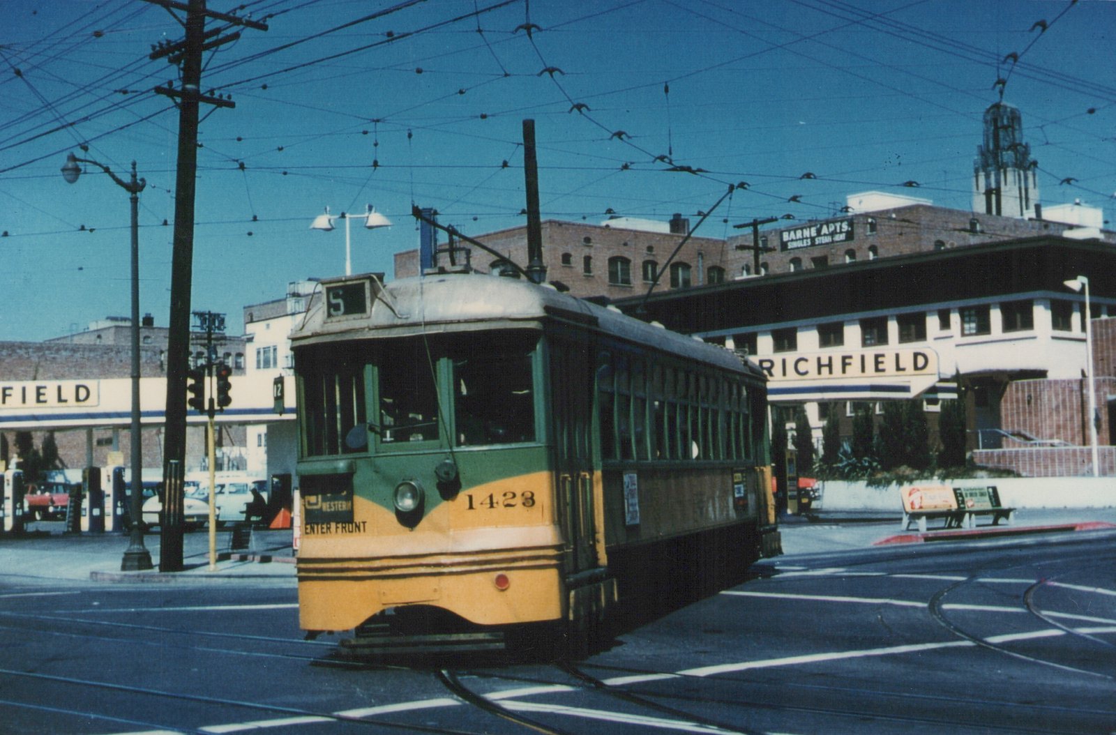 A green and cream streetcar with the number 1423 is stopped at a street intersection on a sunny day, with a Richfield gas station and a building labeled 'BARNE' APTS.' in the background.