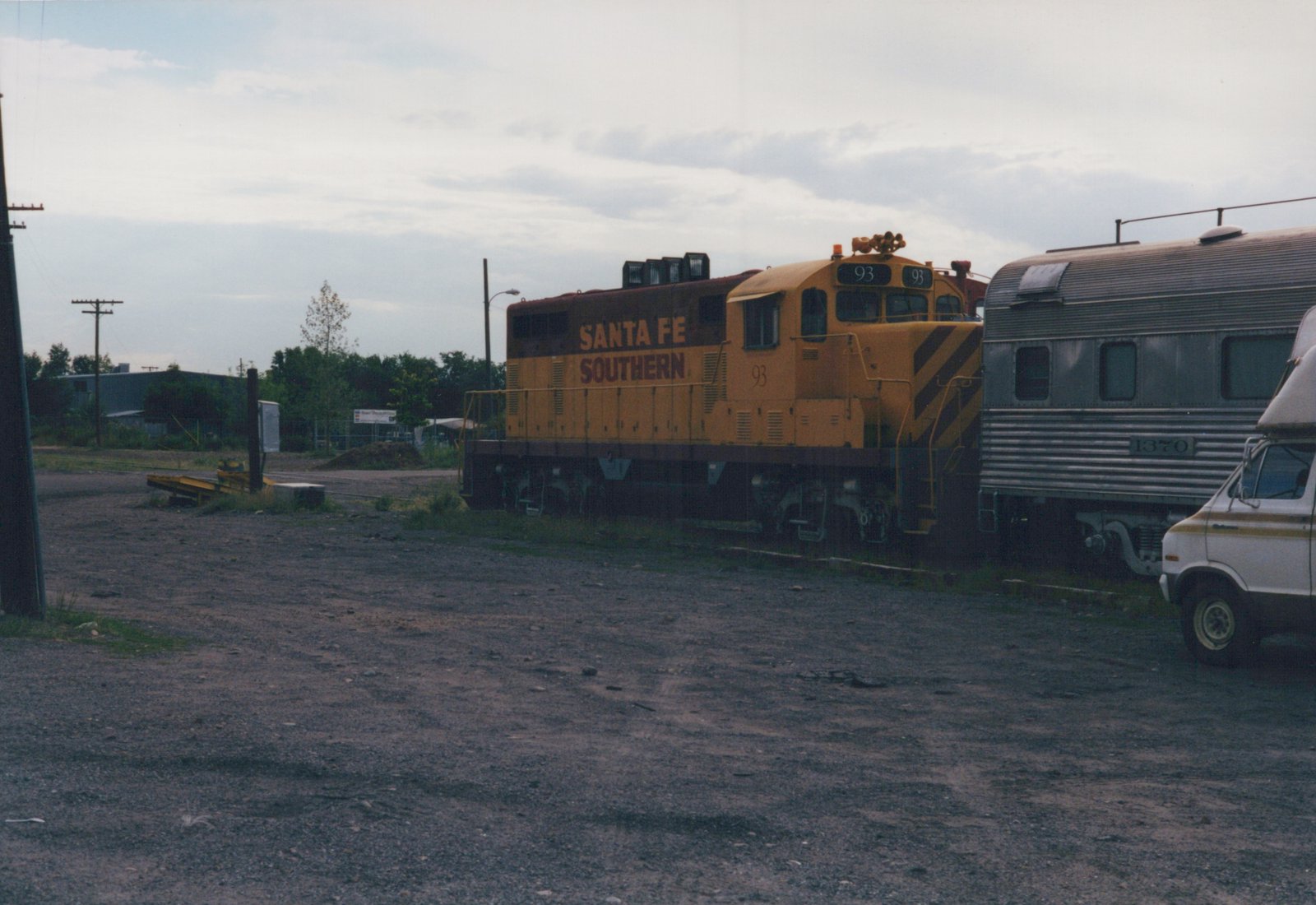 A yellow and red Santa Fe Southern diesel locomotive, number 93, is coupled to a silver stainless steel passenger car, number 1370, in a gravel railroad yard on a cloudy day.