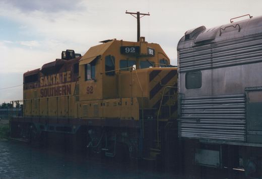 A yellow and red Santa Fe Southern locomotive numbered 92 is coupled to a silver passenger car in a train yard with an overcast sky.