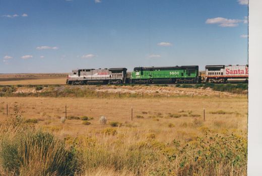 Three diesel locomotives, one silver with LMX lettering, one green with a Burlington Northern logo, and one yellow and red with Santa Fe lettering, are seen traveling on a railroad main line through a grassy field.