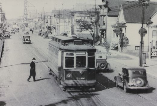 A black and white photo of a 1930s Chicago street scene showing a streetcar on tracks, several old cars, and a person crossing the road, with a Shell gas station and a Capitol sign in the background.