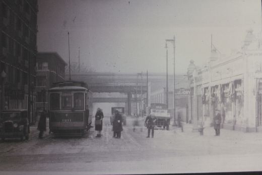 A black and white photo shows a vintage streetcar numbered 1211 on a snowy city street with people, old cars, and storefronts, including a 'MEATS' shop and a 'HARD LAUNDRY'.
