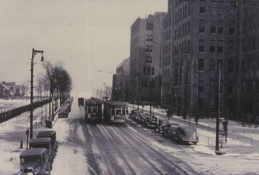 A black and white photo of two vintage streetcars on a snowy city street lined with parked cars and large stone buildings.
