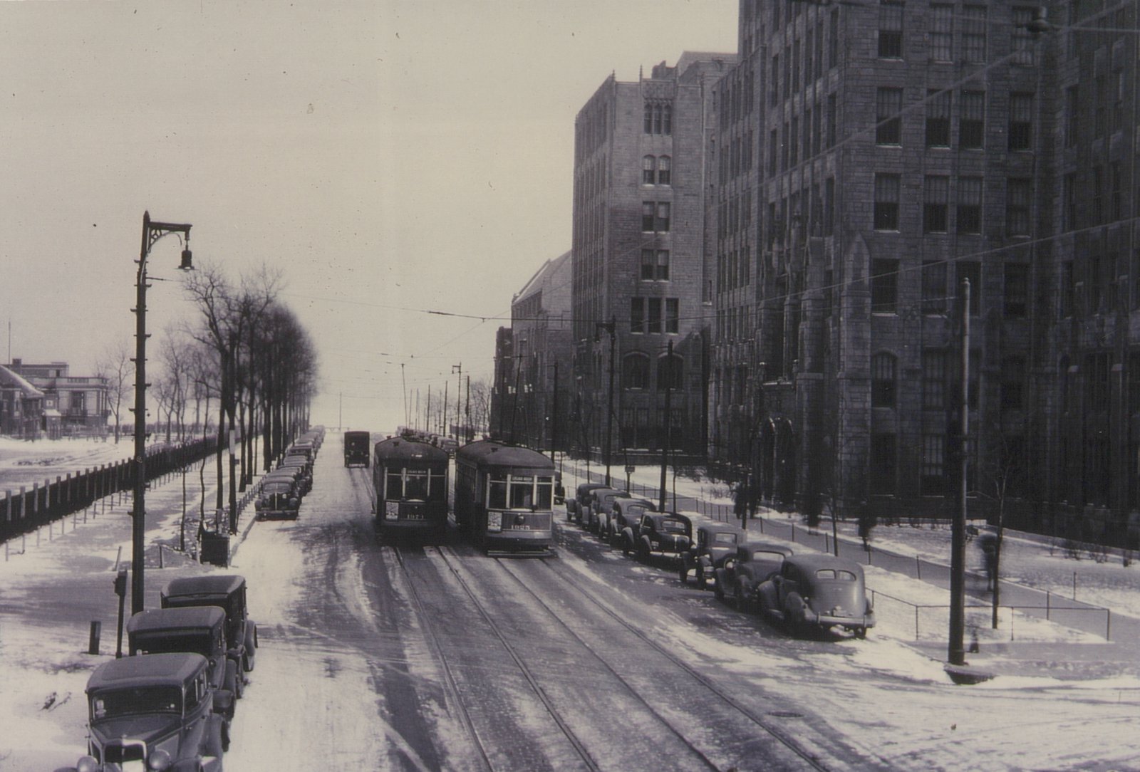 A black and white photo of two vintage streetcars on a snowy city street lined with parked cars and large stone buildings.
