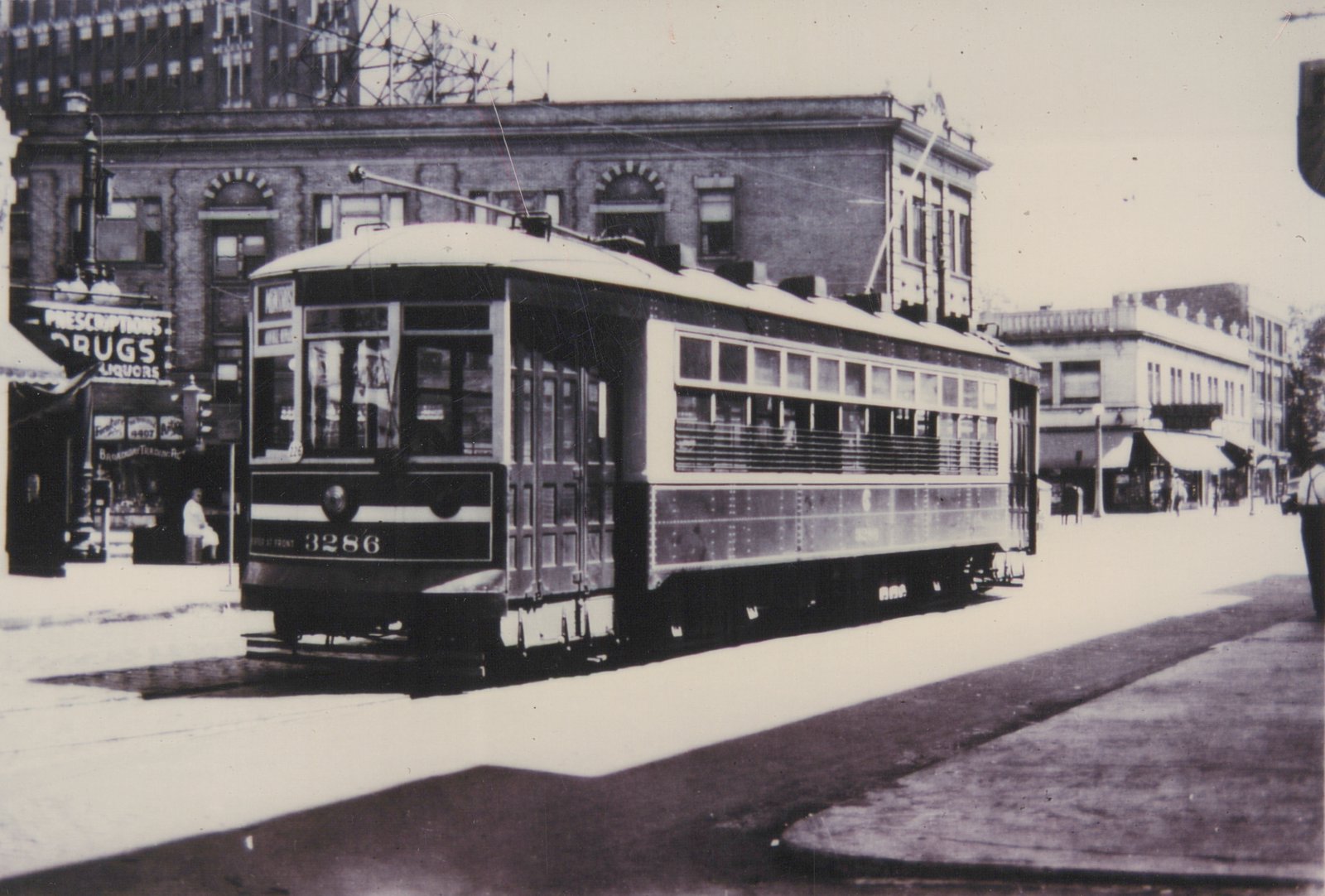 A black-and-white photo of a vintage streetcar, number 3286, on a city street with storefronts and pedestrians visible.