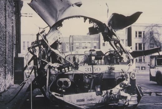 A wrecked and burned bus with the number 3150 on its front, sitting on a city street next to a brick building, with another bus visible in the background.