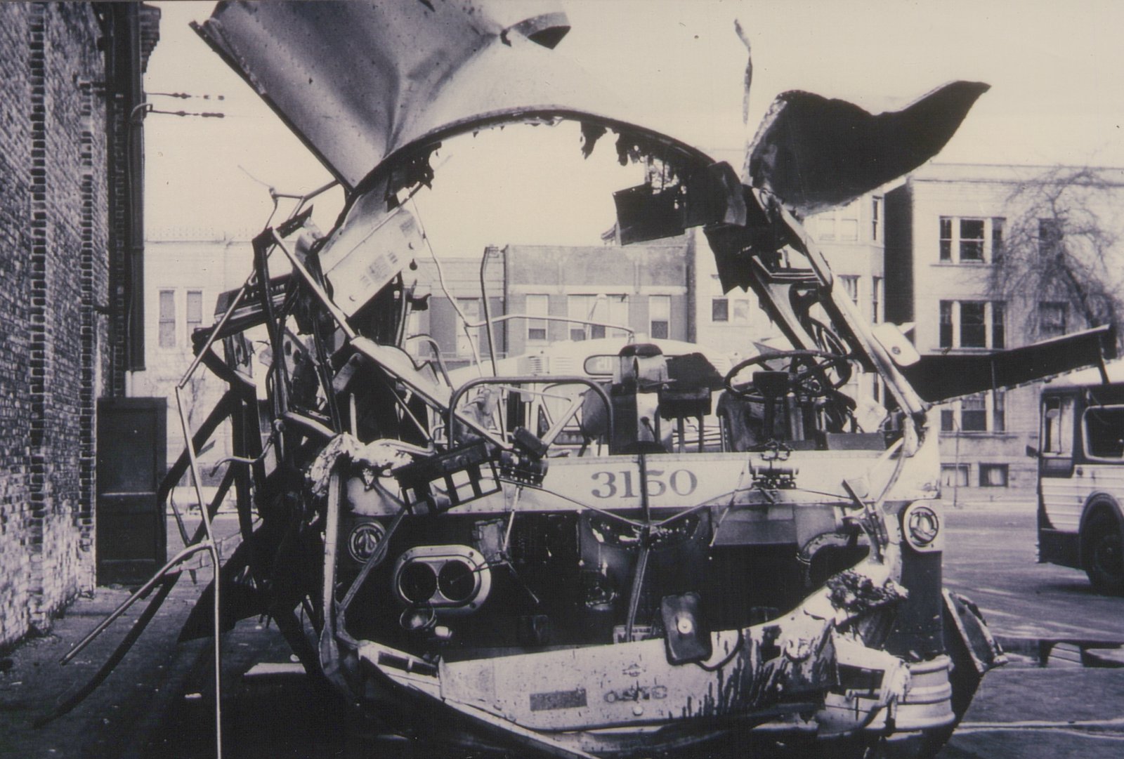 A wrecked and burned bus with the number 3150 on its front, sitting on a city street next to a brick building, with another bus visible in the background.