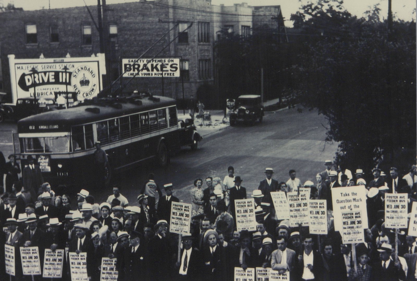 A black and white photo shows a Chicago trolleybus on a city street, with a large crowd of people holding signs in front of it, protesting a bus route change.