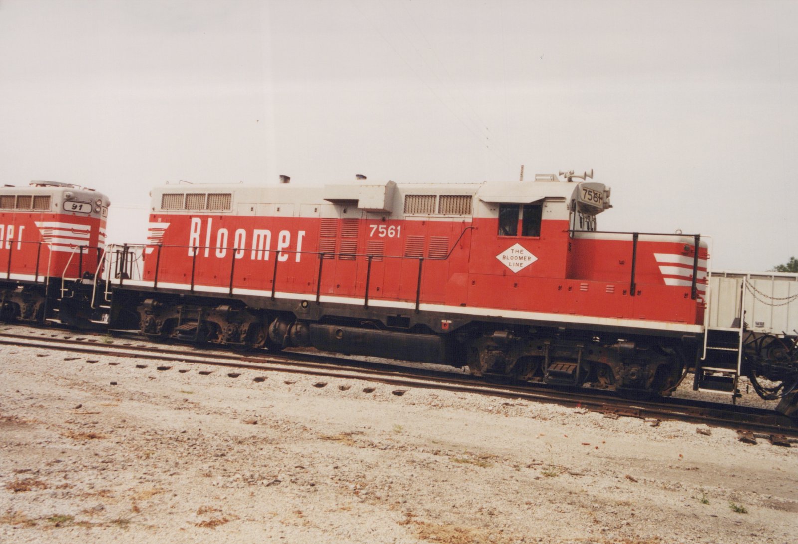Side view of two red and white Bloomer Line diesel locomotives, 7561 and 91, parked on parallel tracks in a gravel yard under an overcast sky.