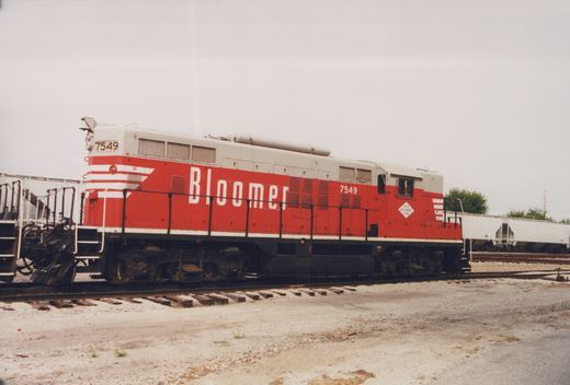 A side view of a red and white diesel locomotive with the number 7549 and 'Bloomer' lettering, parked on tracks in a gravel yard.