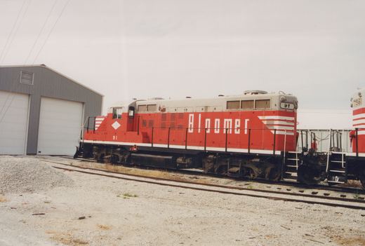 A red and white diesel locomotive with the word 'Bloomer' and number 91 on its side sits on tracks in a gravel yard next to a gray building, with a second red and white locomotive numbered 7561 visible behind it.