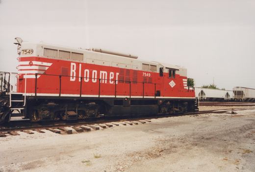 A red and white diesel locomotive with 'Bloomer' lettering and the number 7549, parked on tracks in a gravel yard under an overcast sky.