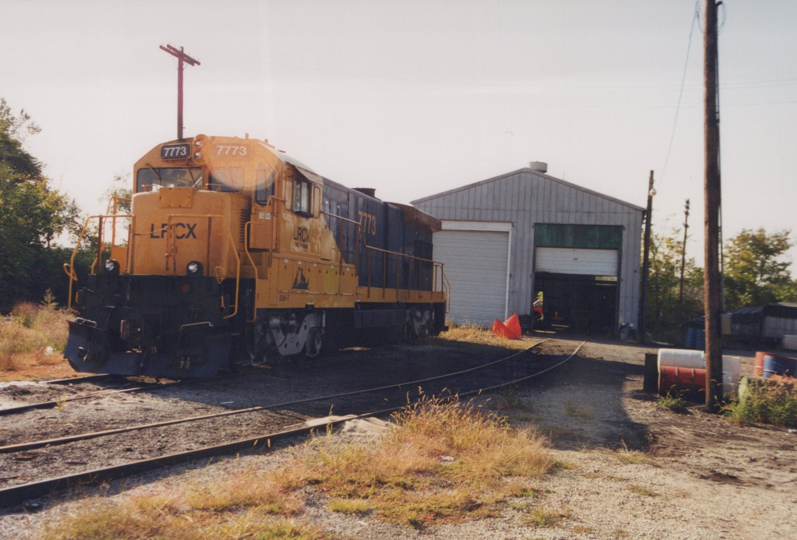 A diesel locomotive painted yellow and blue, numbered 7773 and lettered LRCX, sits on a track in front of a metal building with a person visible in the doorway.