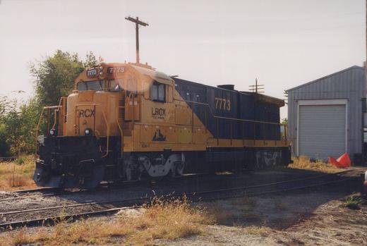 A yellow and black diesel locomotive with the number 7773 and LRCX lettering is parked on tracks in a yard with a building and overgrown grass nearby.
